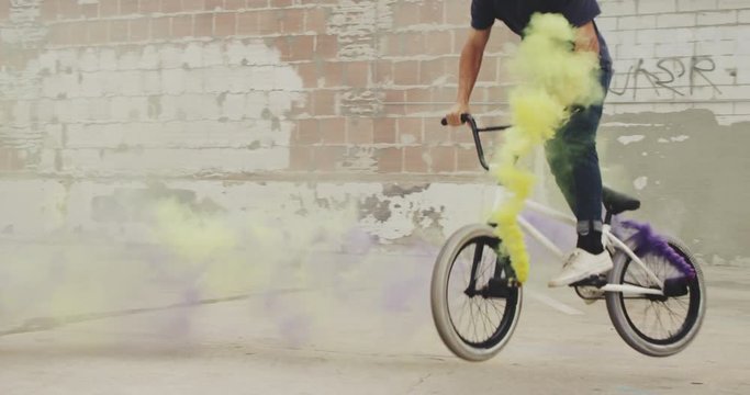 Close up of extreme BMX biker doing tail whip with purple and yellow colored smoke grenade trick in urban environment