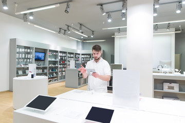 Naklejka na ściany i meble Man buyer buys a tablet in an electronics store. A handsome man stands in a technology store with a tablet in his hand. Purchase a gadget.