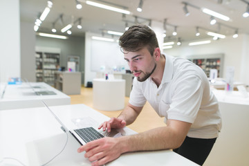 Obraz na płótnie Canvas Portrait of a handsome man using a notebook in a modern technology store with a light interior. The buyer tests the laptop in the electronics store