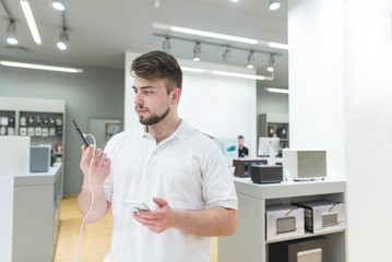 Buyer is in the electronics store in the mobile department with a smartphone in his hands. A handsome man chooses a smartphone in a modern technology store