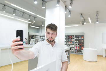 Naklejka na ściany i meble Handsome man makes a selfie on a smartphone in an tech store. Camera test when choosing a smartphone. The buyer selects a smartphone in the electronics store.