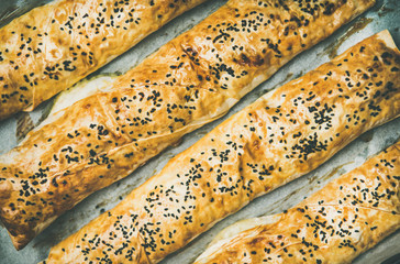 Flat-lay of freshly baked Turkish borek pastry rolls with spinach, feta cheese and black cumin seeds over baking paper, top view. Traditional East Mediterranean cuisine