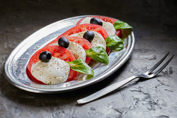 Caprese Salad. Tomato and Mozzarella slices, Basil and olives is on a plate. Dark background.