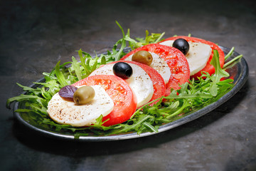 Mozzarella with sliced tomatoes, Basil and olives is on arugula in a plate. Dark background.