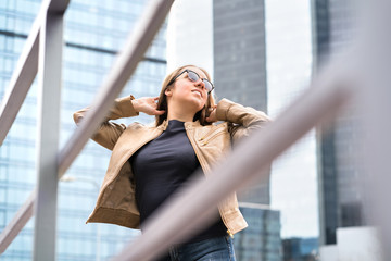 Independent, confident and powerful woman in city. Happy healthy model with sunglasses and good fashion style. Big, tall and high modern office buildings. Lady posing hands in hair.