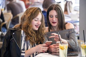 Two young and trendy hipster woman are working in a cafe bar, checking something on a phone. Lifestyle conception.