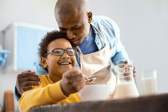 Tender love. Caring young father hugging his little son at breakfast while pouring milk into his bowl of cereals