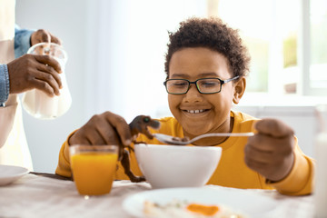 Fototapeta na wymiar Power of imagination. Upbeat pre-teen boy feeding his toy dinosaur with cereals and smiling happily while playing during breakfast