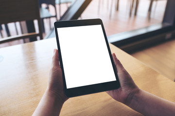 Mockup image of hands holding black tablet pc with blank white desktop screen on wooden table