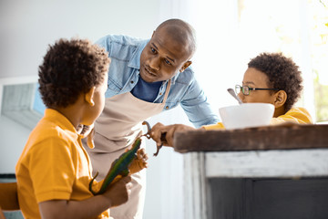 Keep calm. Caring young father asking his sons to behave themselves while they playing with toy dinosaurs too loudly during breakfast