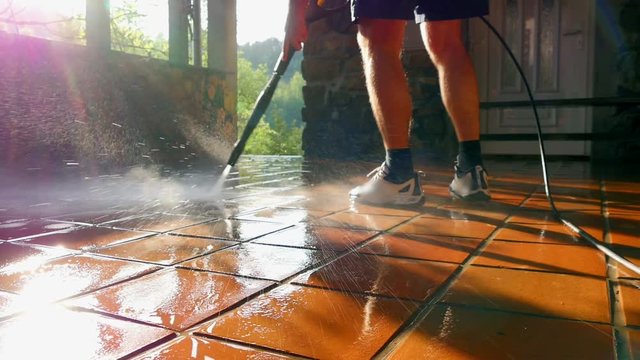 Male in shorts cleans outdoor terrace with high pressure washer. Water spraying in beautiful sunset making rainbow colored lens flares.