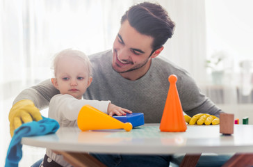 Handsome man in yellow gloves cleaning table while his little child playing toys, modern father