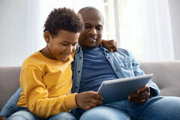 Fototapeta premium Interesting videos. Cheerful pre-teen boy hugging his father and watching videos together with him while relaxing on sofa