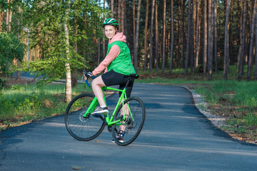 Naklejka premium Beautiful smiling blond girl is riding the bicycle in the park. Green vest, bicycle and green helmet. Doing sports.