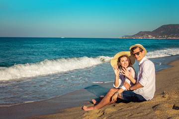 Fototapeta premium Young happy couple sitting on tropical beach and waving at camera. Travellers chilling and relaxing