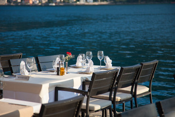 Romantic dinner by sea restaurant with beautiful view on mountains. Served table with white and beige tableclothes, wineglasses of ice champagne, red flowers.