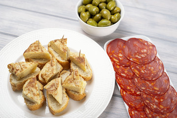 Plate with omelette caps with bread next to plate with chorizo and a bowl with olives on wooden table. Typical spanish food.