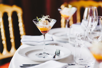 Wedding table decorated with white plates, glasses for wine and cocktail in spacious hall. Elegant dinner table against cozy interior. Anniversary celebration