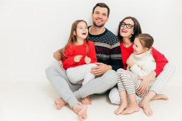 Studio shot of happy family members sit on floor with bare feet, embrace each other, have joyful expression, laugh as hear positive news or funny story, isolated on white background. Parents, children