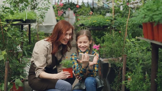 Smiling young woman and her cute child are using smartphone, touching screen and laughing inside greenhouse. Modern technology, happy family and people concept.
