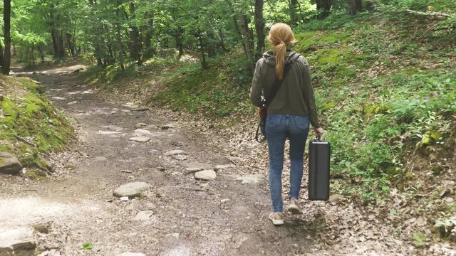 girl, walks through the woods with a heavy black suitcase. 4k, slow-motion shooting, steadicam shot.