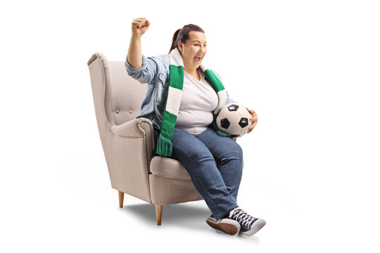 Female soccer fan with a scarf and a football sitting in an armchair and cheering