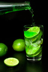Glass of cold green lemonade with ice and fresh ripe green limes on black