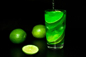 Glass of cold drink with ice and fresh ripe slice green limes on black background.