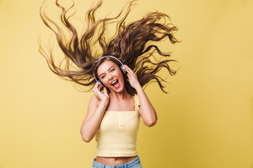 Image of amusing woman 20s singing and having fun with shaking hair while listening to music via headphones, isolated over yellow background