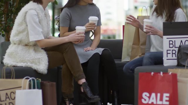 Tilt up shot of three young women sitting on sofa in shopping mall and talking to each other while drinking takeaway coffee after buying clothes