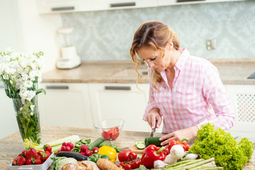 smiling woman in a shirt is slicing cucumber on a kitchen table with vegetables