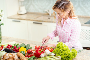 smiling woman in a shirt is slicing tomatoes on a kitchen table with vegetables