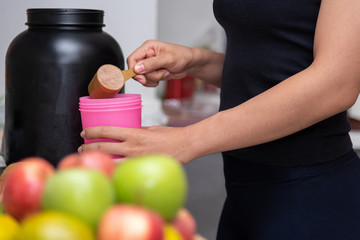 Healthy women preparing a whey protein after doing weight training in the kitchen with fresh fruits as a blurred foreground.