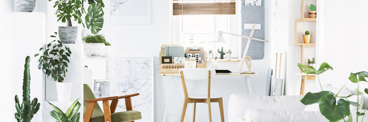 Bright home office interior with appliances and wooden lamp on desk, window with blinds and fresh green plants