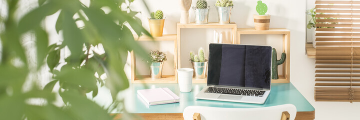 Green plant in blurred foreground in the photo of computer for remote work placed on small desk with calendar and coffee cup