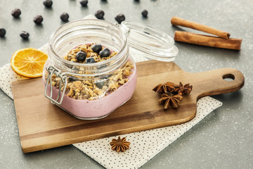 Obraz na płótnie Canvas Pink, fruit yogurt with granola and blueberries in a jar on a wooden cutting board and cinnamon spice on a gray kitchen counter