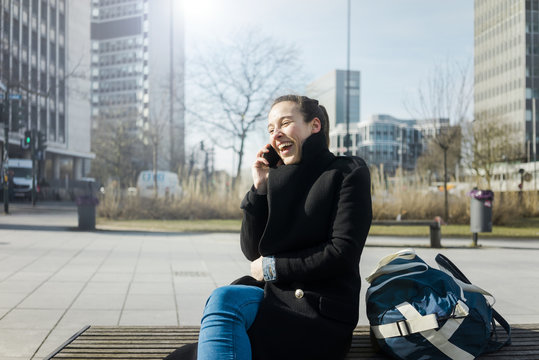 Germany, Essen, laughing young woman on the phone sitting on bench outdoors