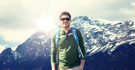 travel, tourism and people concept - happy young man in sunglasses with backpack over alps mountains background