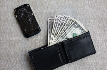 Crashed black smartphone and money in leather wallet to buy a new one. Dollars and broken phone on the rustic tablecloth. Time to get a loan for electronics.