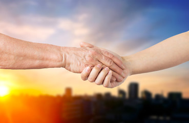 people, old age and generation concept - close up of senior and young woman holding hands over city sunset background