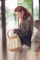 beautiful young woman waiting at a bus stop looking in her hand bag, purse, autumn mood colors
