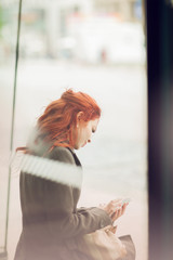 beautiful young woman waiting at a bus stop looking at her cell phone, autumn mood colors