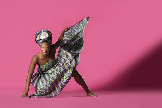 Beautiful African Black girl wearing traditional colorful African outfit does a dramatic dance move against a colorful pink background