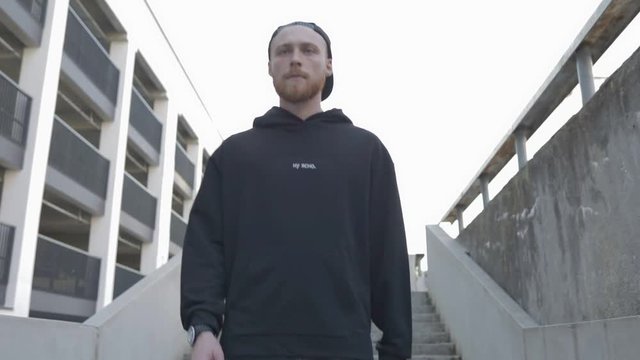 A handsome serious guy in a black sweatshirt and a cap goes down the stairs. City urban style. Slow motion video shooting by handheld gimbal