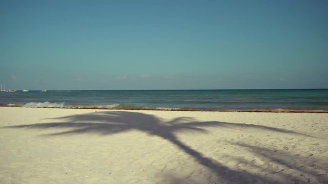 View of tropical beach on sunset. Shadows of palm tree fronds fluttering on textured sand beach. Parasailing boats on pier. Turquoise water of the Caribbean Sea. Riviera Maya Mexico.