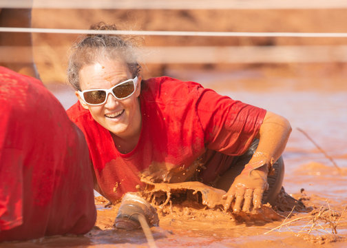 Woman in a red shirt and white sunglasses smiling while crawling through a mud obstacle during a mud run 