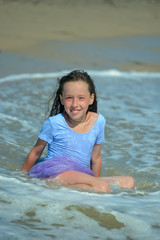 A charming little girl sits on the ocean on a Sunny day in a purple swimsuit, and a wave runs over her. She looks at the camera and smiles. Vertical orientation of the image