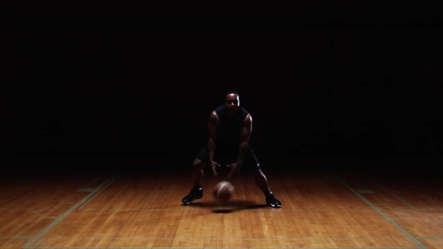 A basketball player doing tricks and dribbling through his legs on a dark court
