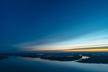 Beautiful river with big island with trees under predawn sky. Bright yellow stripe in picturesque cloudy sky. Early blue sky reflected in water. Colorful morning atmospheric image of majestic nature.