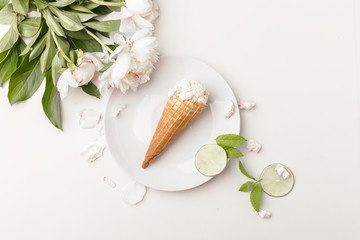 Flatlay of sweet cones of cream ice cream on white plate, slices of lime, mint leaves and white peonies on white background, top view, copy space